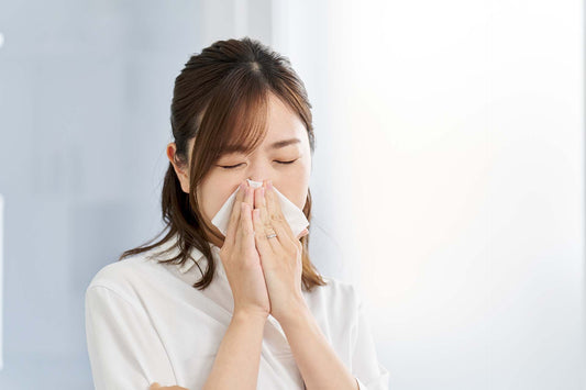 Woman in white holding a tissue to her nose in a bright room, a sign of nasal allergy symptoms.