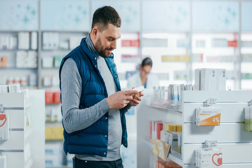 Man in a blue vest reading a product box while shopping a pharmacy allergy-medicine aisle