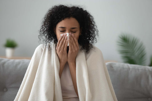 Woman wrapped in a blanket on a couch blowing her nose into a tissue, with plants in the background