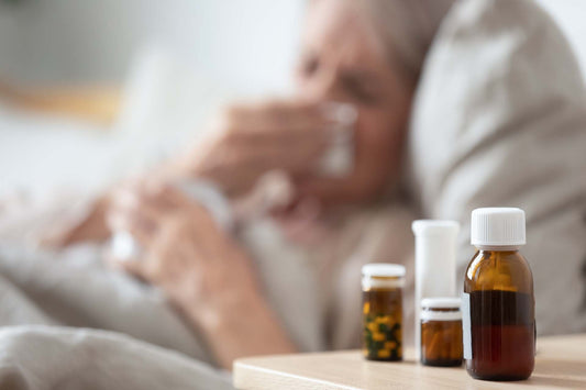 Sick person resting in bed blowing their nose, with medication bottles on the bedside table.
