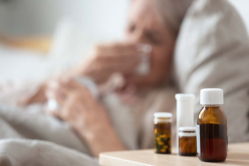 Sick person resting in bed blowing their nose, with medication bottles on the bedside table.