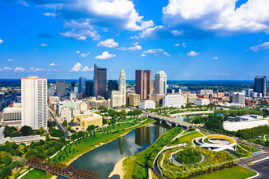 Aerial view of the Columbus, Ohio skyline along a river, with a park, bridges, and a bright cloudy blue sky