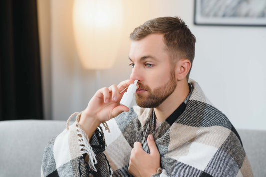 Man wrapped in a plaid blanket on a couch using a nasal spray, a warm lamp glowing behind him