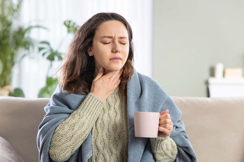 Woman wrapped in a blanket holding a mug and touching her throat, eyes closed, on a couch