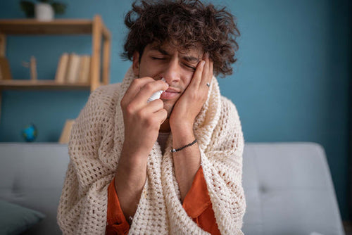 Person holding a nasal spray while wrapped in a blanket with cold or allergy symptoms.