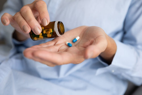 Person tipping capsules from a pill bottle into their open palm, including blue-and-white allergy medication.