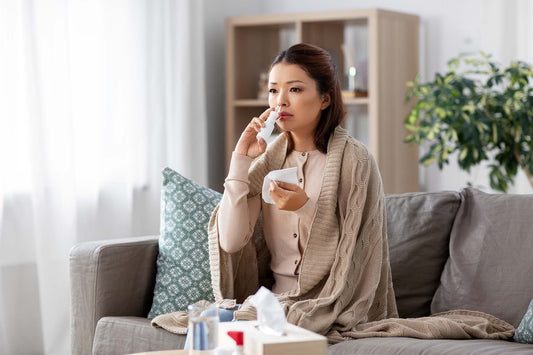 Person using a nasal spray while sitting on a couch at home, wrapped in a cardigan.