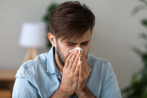 Man in a denim shirt blowing his nose into a tissue at home, a symptom of allergic rhinitis.