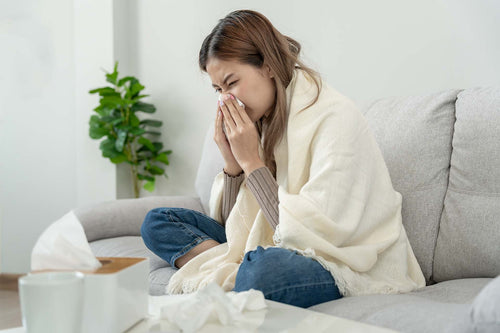 Woman wrapped in a blanket on a couch blowing her nose, with tissues and a mug nearby, a sign of allergy symptoms.
