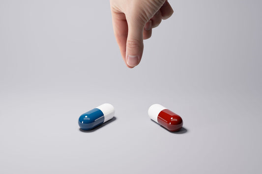 Hand reaching toward two capsule pills, one blue-and-white and one red-and-white, on a gray surface.