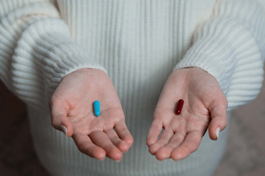 Two open palms each holding a single pill, one blue capsule and one dark red pill.